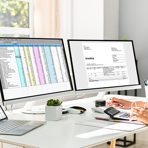 office with two monitors displaying spreadsheets as a person works on online bookkeeping